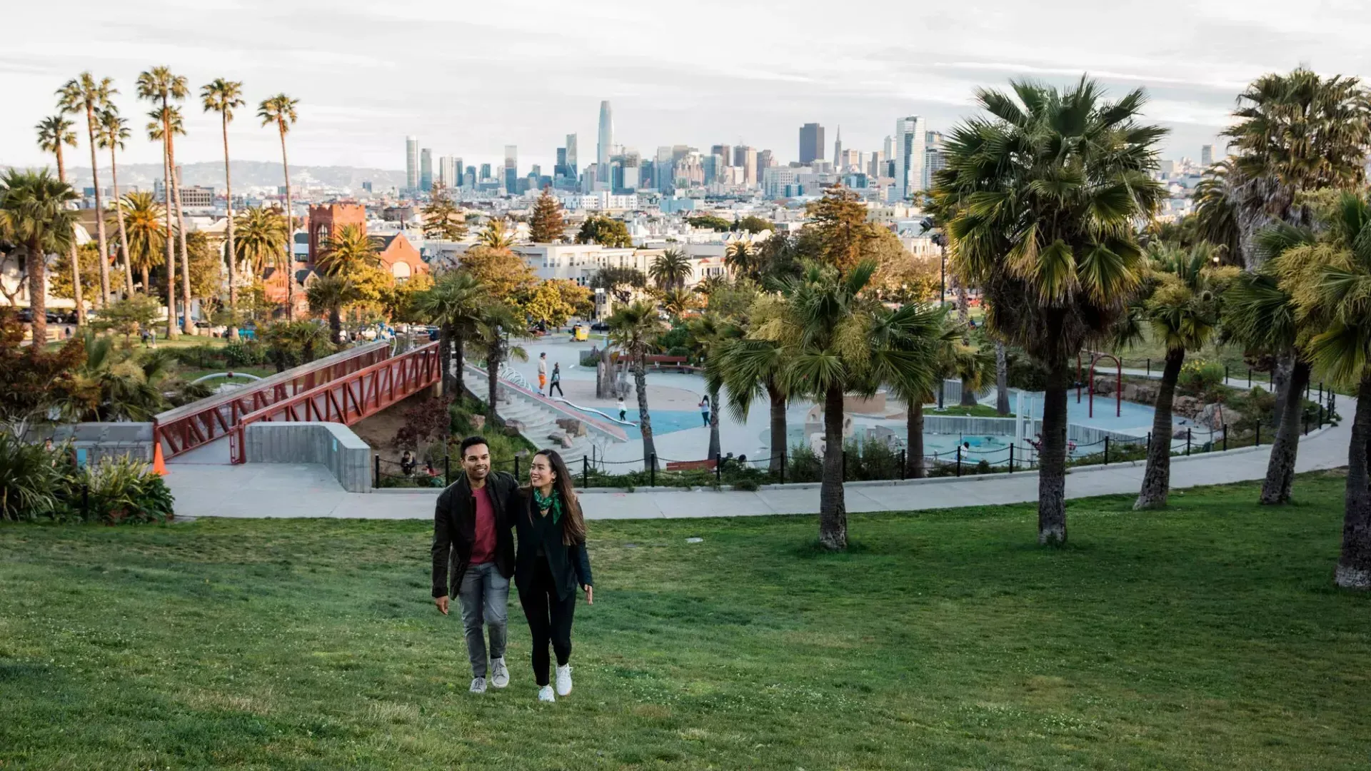 Una pareja camina hacia la cámara con Dolores Park y el horizonte de San Francisco detrás de ellos.